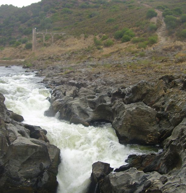 Rio com corredeiras em um vale rochoso, cercado por vegetação e morros ao fundo.
