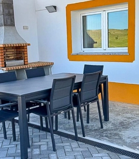 Área externa com churrasqueira, mesa preta com cadeiras, parede branca e janela com moldura laranja.