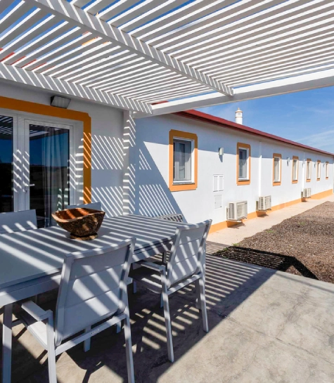 Terraço com mesa e cadeiras sob pérgola, sombra de ripas projetada no chão.