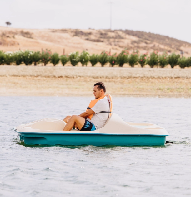 Homem em pedalinho azul e bege, usando colete, em lago sereno.