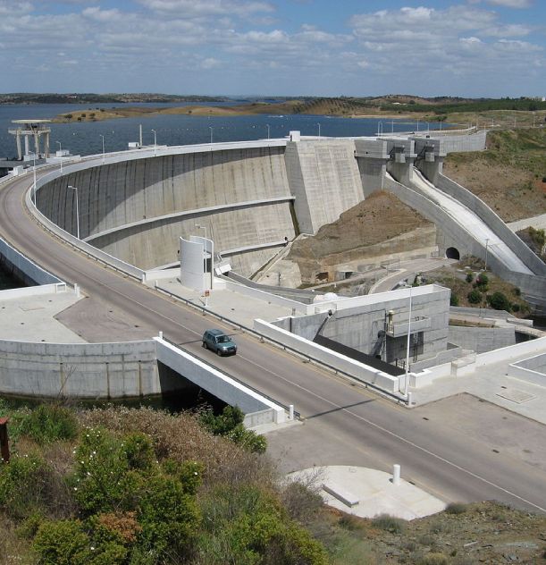 Barragem de concreto com estrada curvada, cercada por água e vegetação. Céu parcialmente nublado.