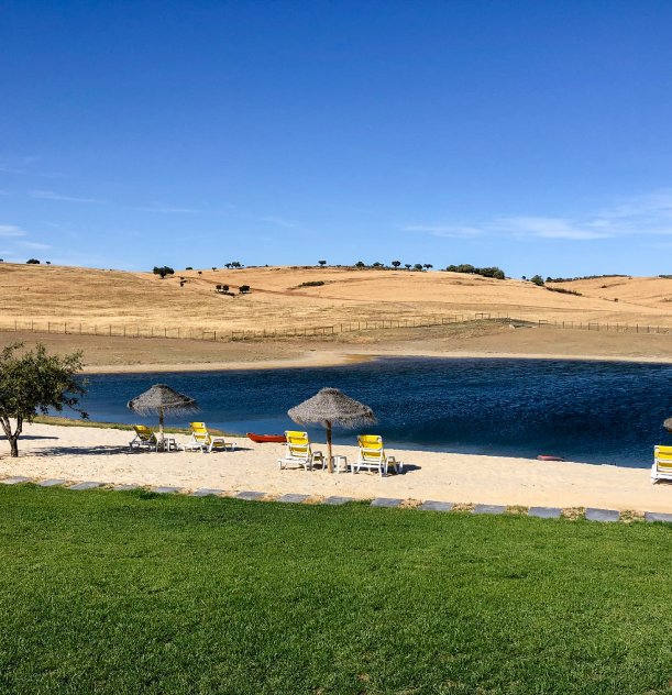 Praia com espreguiçadeiras e guarda-sóis, lago ao fundo, campo seco e céu azul.