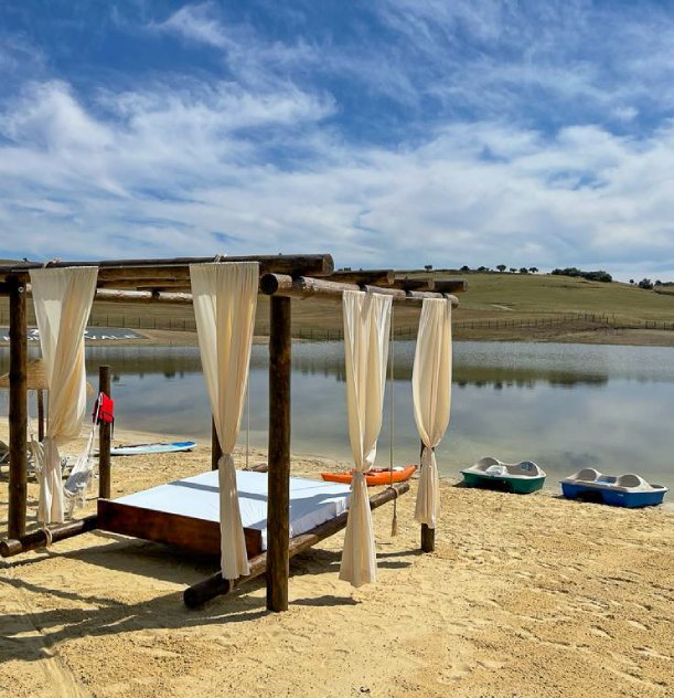 Pérgola de madeira com cortinas brancas à beira do lago, céu azul e barcos ao fundo.