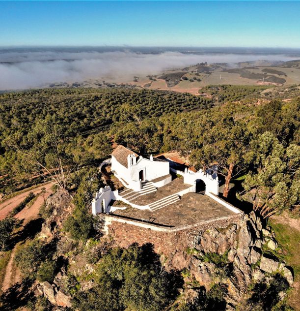 Igreja branca em colina arborizada com paisagem rural ao fundo e céu azul.