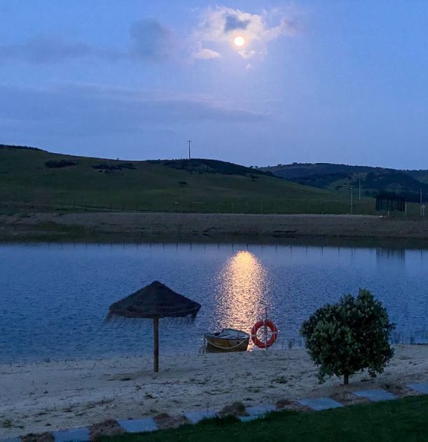 Lago ao entardecer com lua refletida, montanhas ao fundo e guarda-sol de palha na areia.