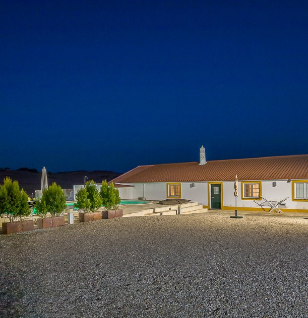 Casa térrea iluminada, com paredes brancas e telhado vermelho, ao anoitecer.