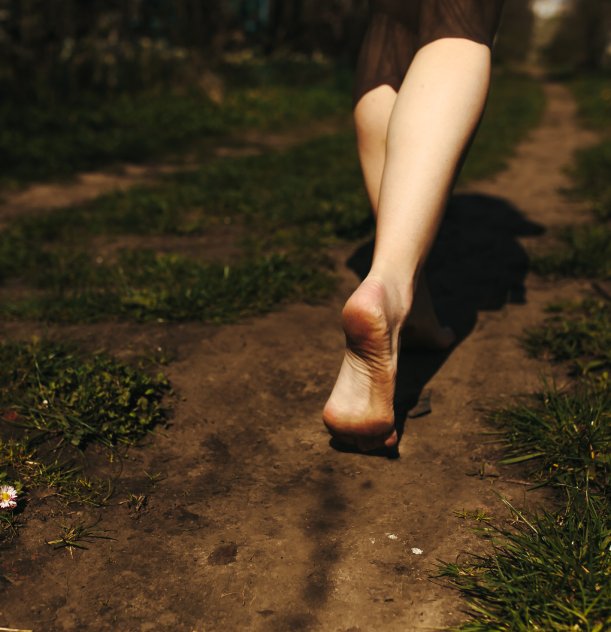 Pessoa caminhando descalça em um caminho de terra cercado por grama.