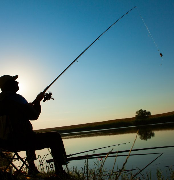 Pescador sentado à beira de um lago, ao entardecer, segurando uma vara de pesca contra o céu.