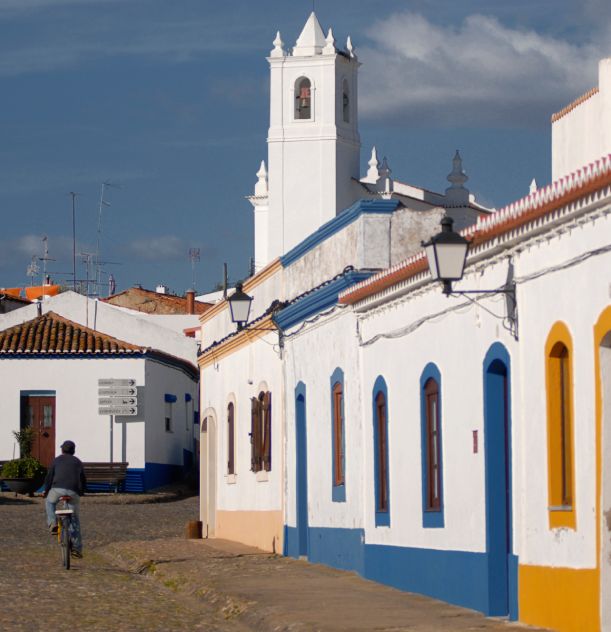 Rua com casas coloridas e uma pessoa de bicicleta, torre de igreja ao fundo.