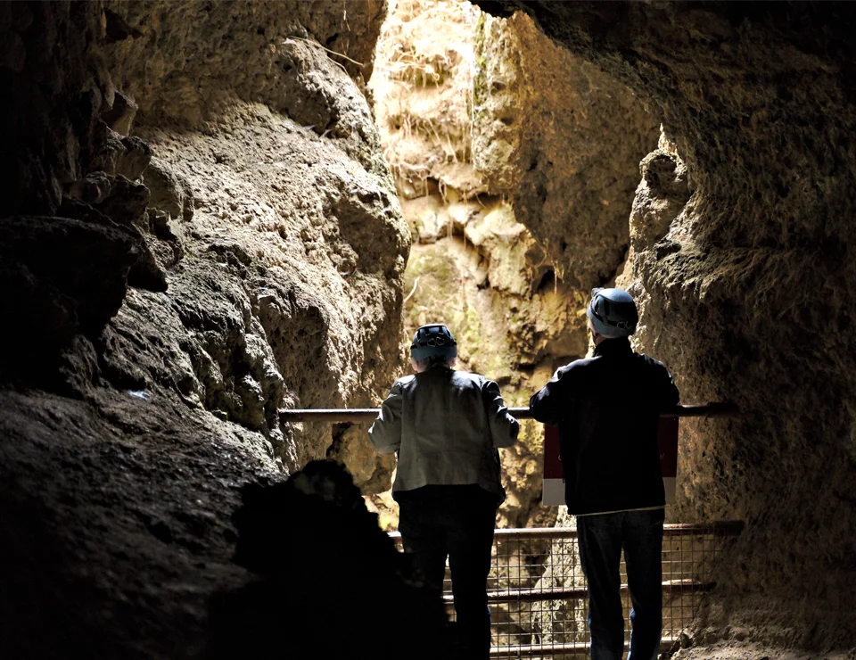 Dos personas con cascos miran hacia la entrada iluminada de una cueva rocosa.