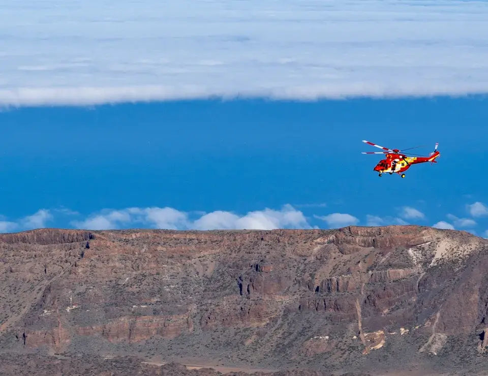 Helicóptero volando sobre un paisaje montañoso con cielo despejado y nubes al fondo.