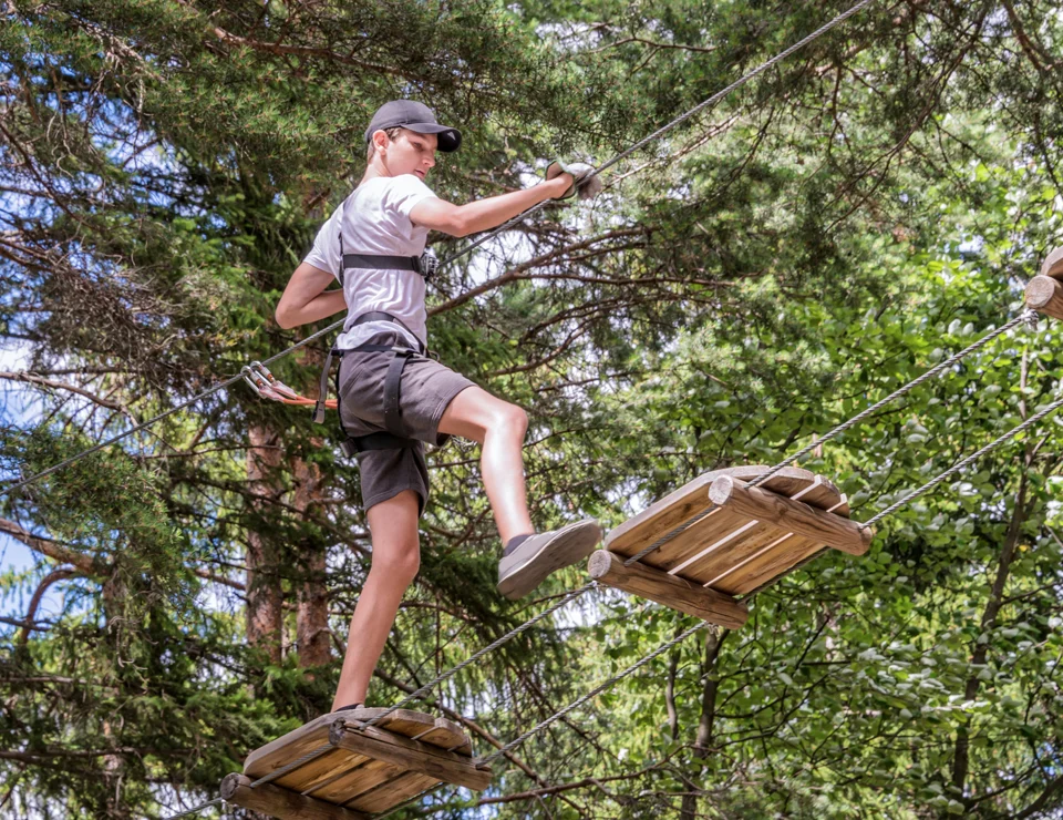 Persona cruzando un puente colgante de madera entre árboles en un parque de aventuras.
