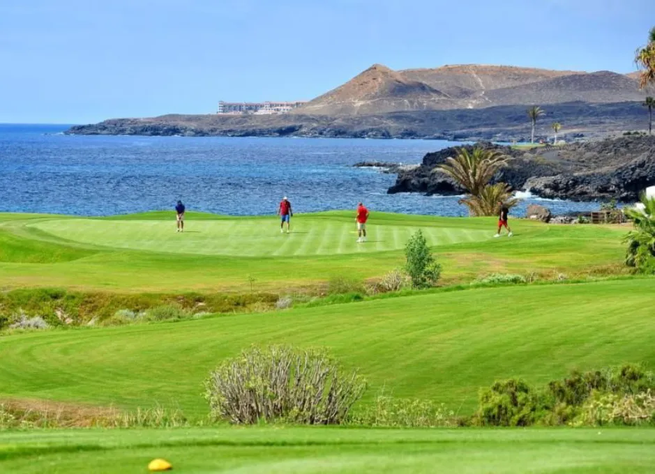 Jugadores de golf en un campo junto al mar, con montañas al fondo.