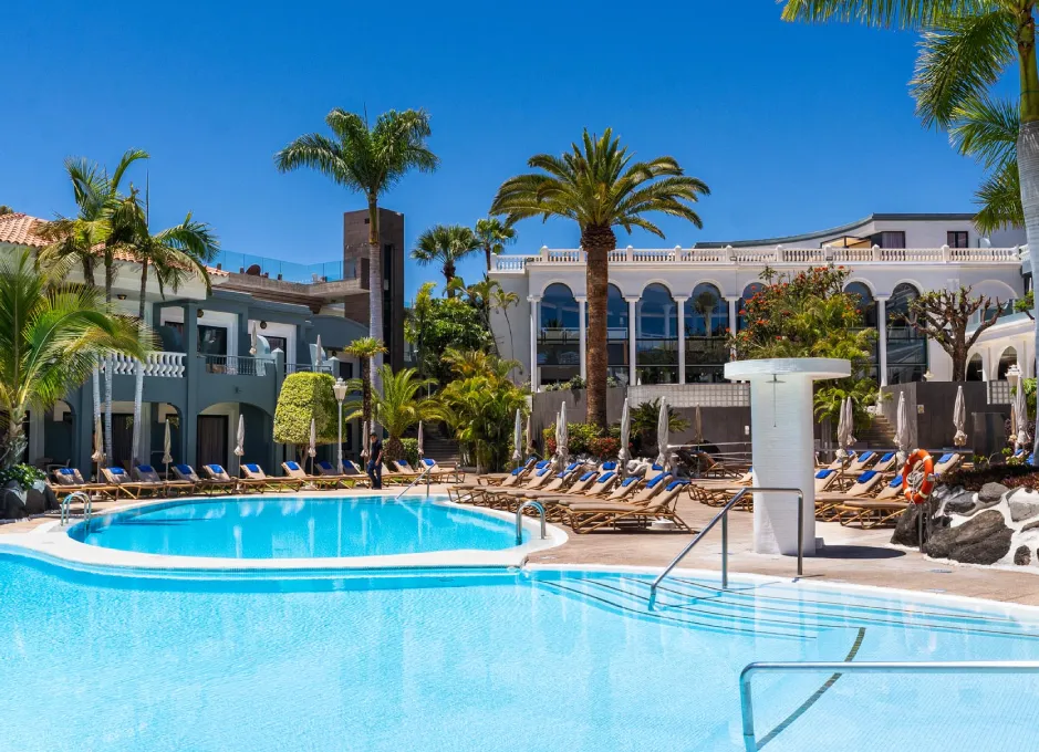 Piscina rodeada de palmeras y tumbonas, con edificios de estilo mediterráneo al fondo.