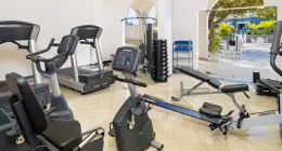 Gimnasio con bicicletas, caminadoras, remo y pesas. Ambiente luminoso con vista al exterior.