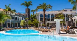 Piscina en un hotel con palmeras, tumbonas y edificio de arquitectura mediterránea.