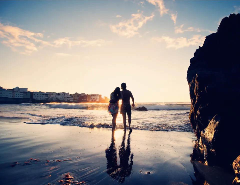 Pareja caminando por la playa al atardecer, con reflejo en el agua y edificios al fondo.