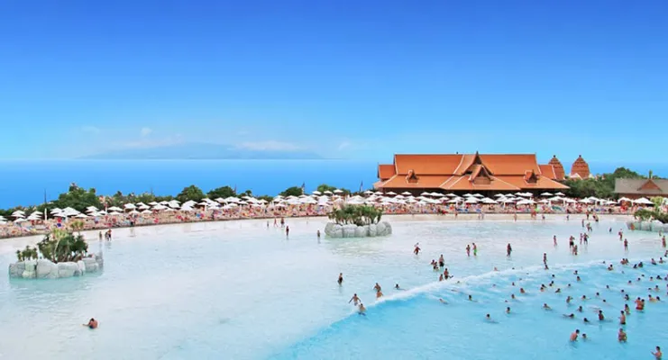 Piscina grande con bañistas, edificaciones al fondo y cielo azul.