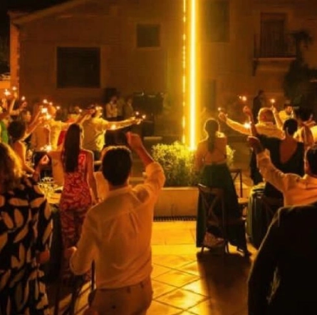 Personas levantando copas, rodeando una estructura luminosa en una celebración nocturna al aire libre.