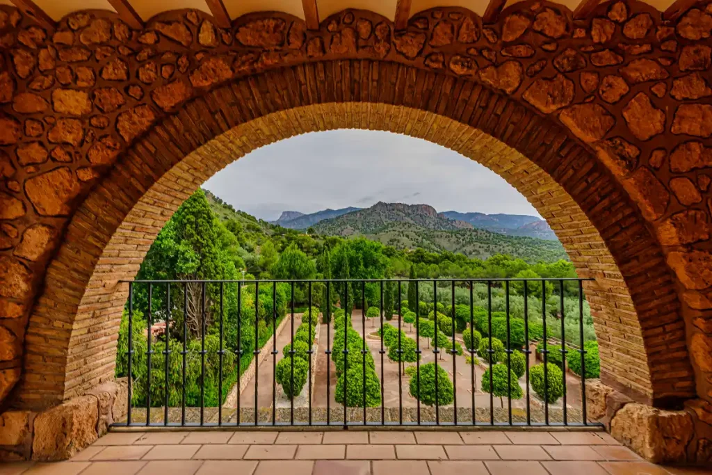 Arco de piedra en un balcón con vistas a un jardín y montañas al fondo.