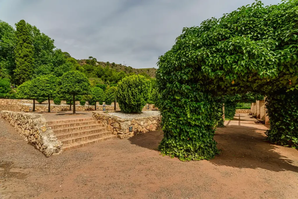 Entrada a un jardín con arcos de enredaderas, árboles y un sendero de piedra.
