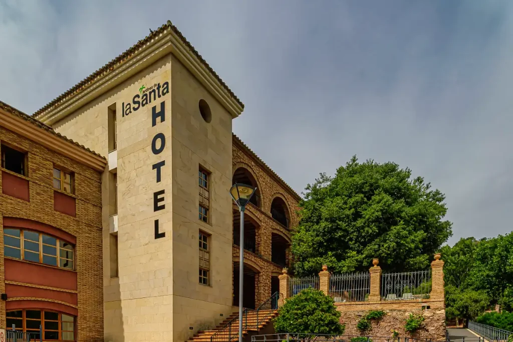 Fachada de un hotel con un árbol grande al lado y cielo despejado de fondo.
