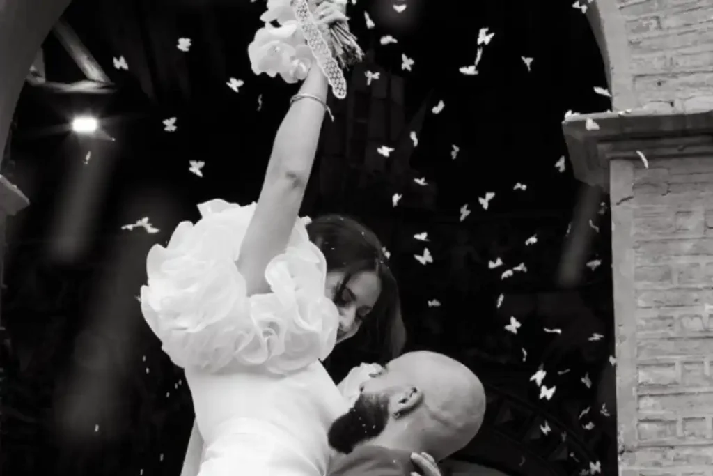 Pareja bailando en boda, bajo lluvia de pétalos. Ella levanta un ramo con sonrisa. Blanco y negro.