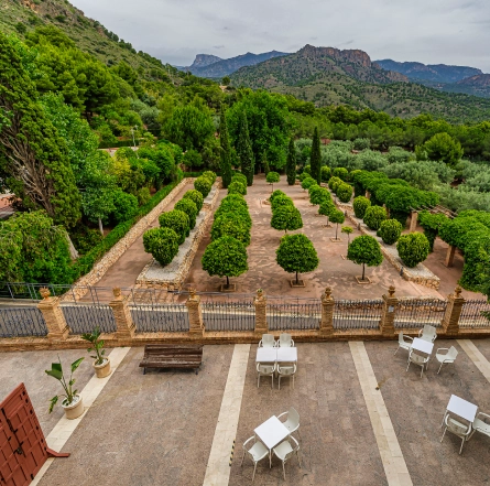 Patio con mesas y sillas, huerto, árboles y montañas al fondo bajo un cielo nublado.