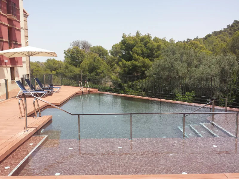 Piscina al aire libre rodeada de sillas, sombrilla y vegetación.