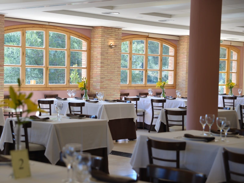 Restaurante elegante con mesas cubiertas por manteles blancos y ventanas grandes con vista a árboles.