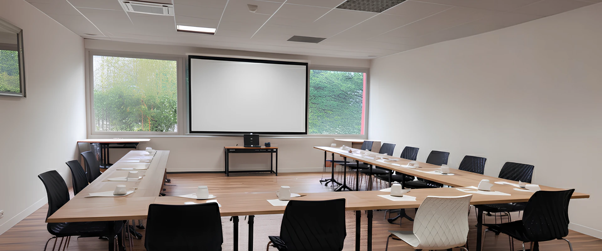 Sala de reuniones vac&iacute;a con mesas en forma de U, sillas negras y pantalla de proyecci&oacute;n.