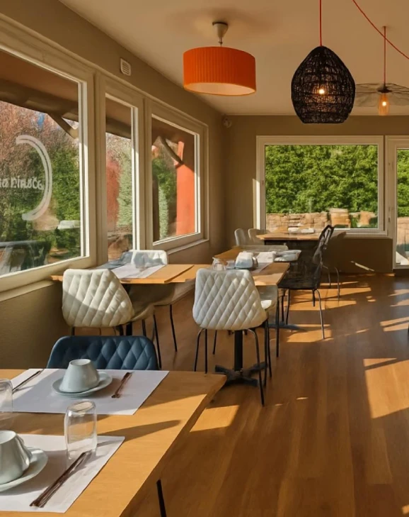 Restaurante luminoso con mesas junto a grandes ventanas, sillas acolchadas, y l&aacute;mparas colgantes.