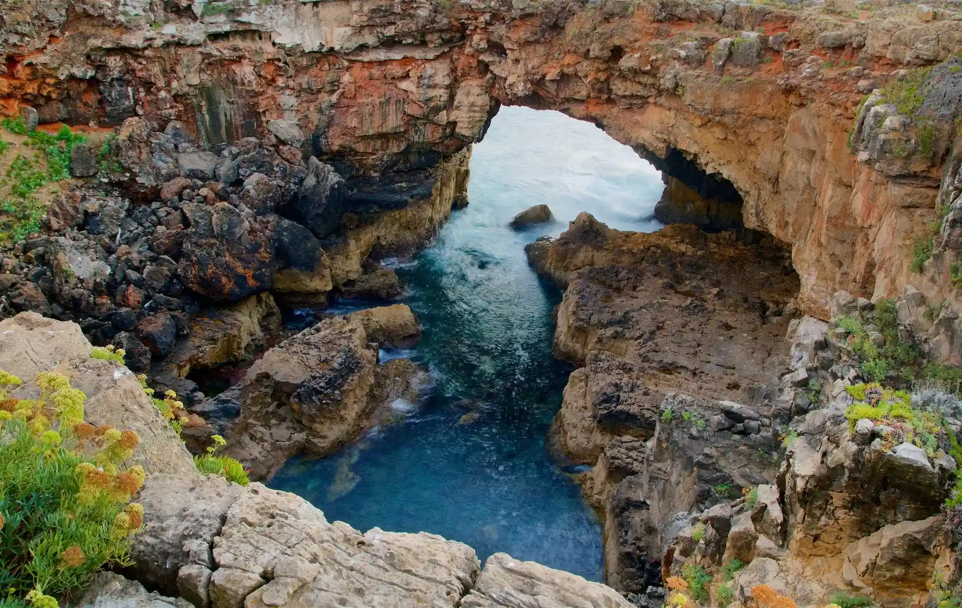 Rocky coastal arch with blue water beneath and green foliage surrounding the edges.