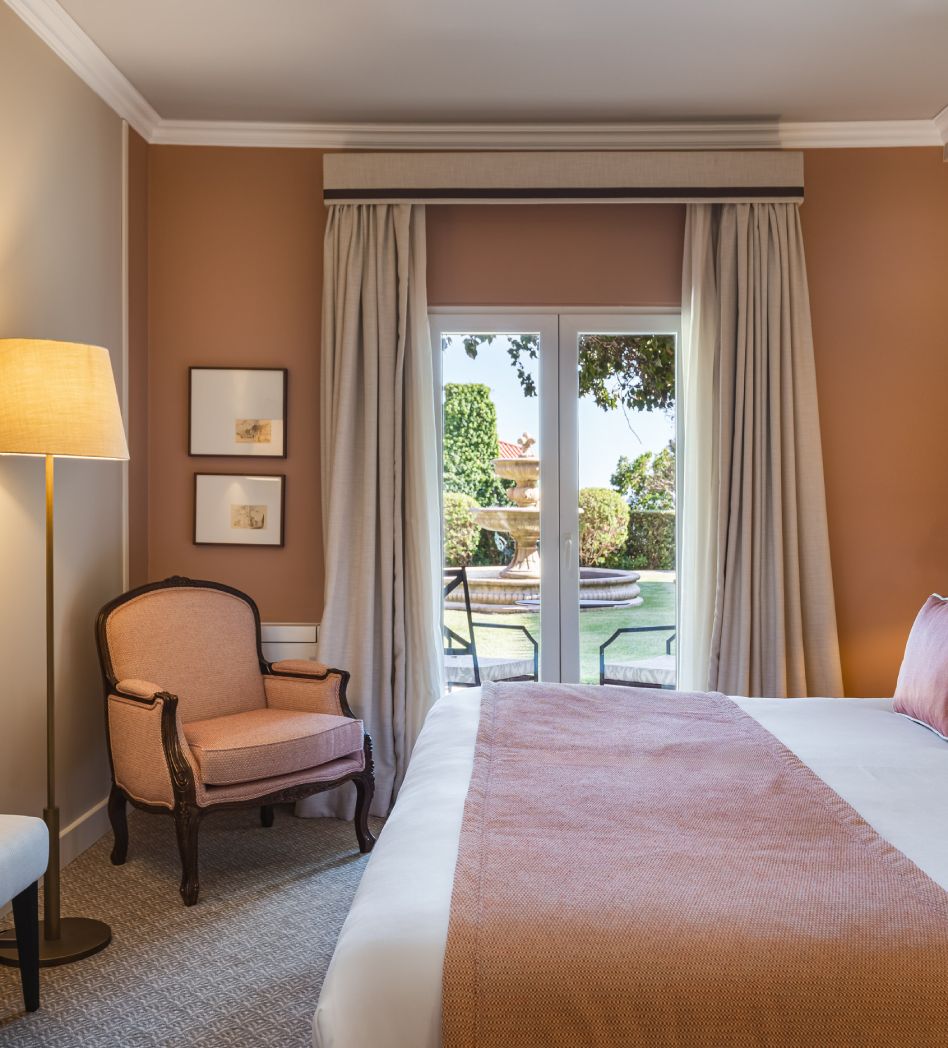 Cozy bedroom with a peach armchair, bed, and window view of garden and fountain.