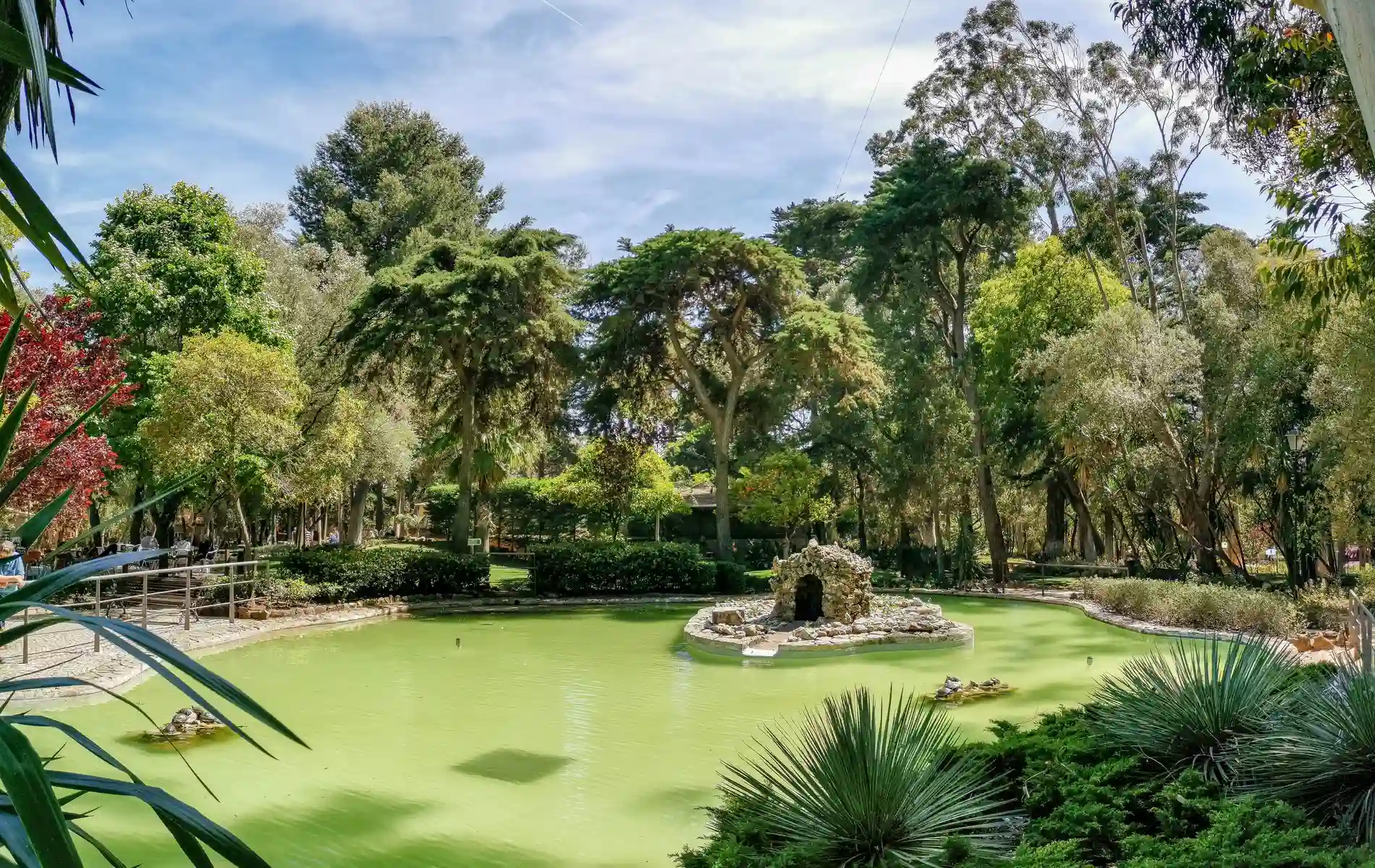 Lush park with trees, a small pond, and rocky structure under a clear blue sky.