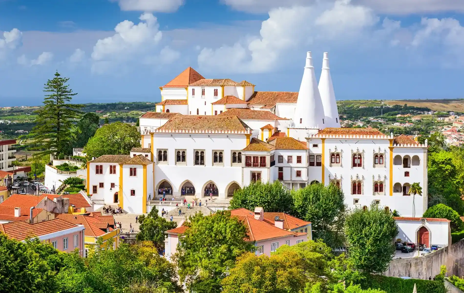 White palace with conical chimneys, surrounded by greenery, under a blue sky with clouds.