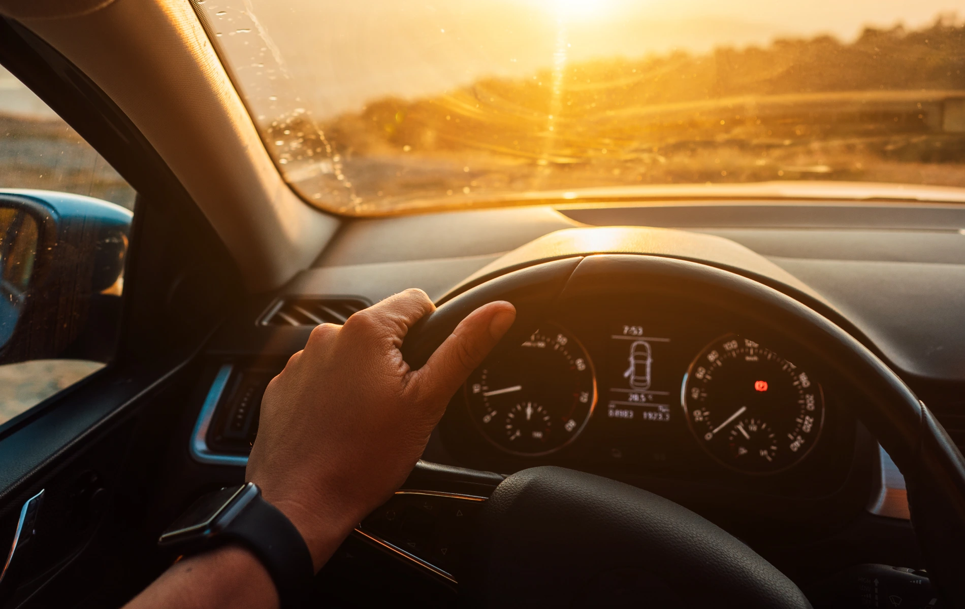 Hand on car steering wheel at sunset, dashboard visible.