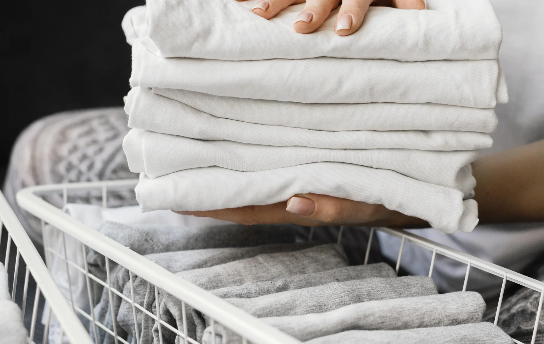 Hands holding a stack of neatly folded white shirts above a basket of folded clothes.
