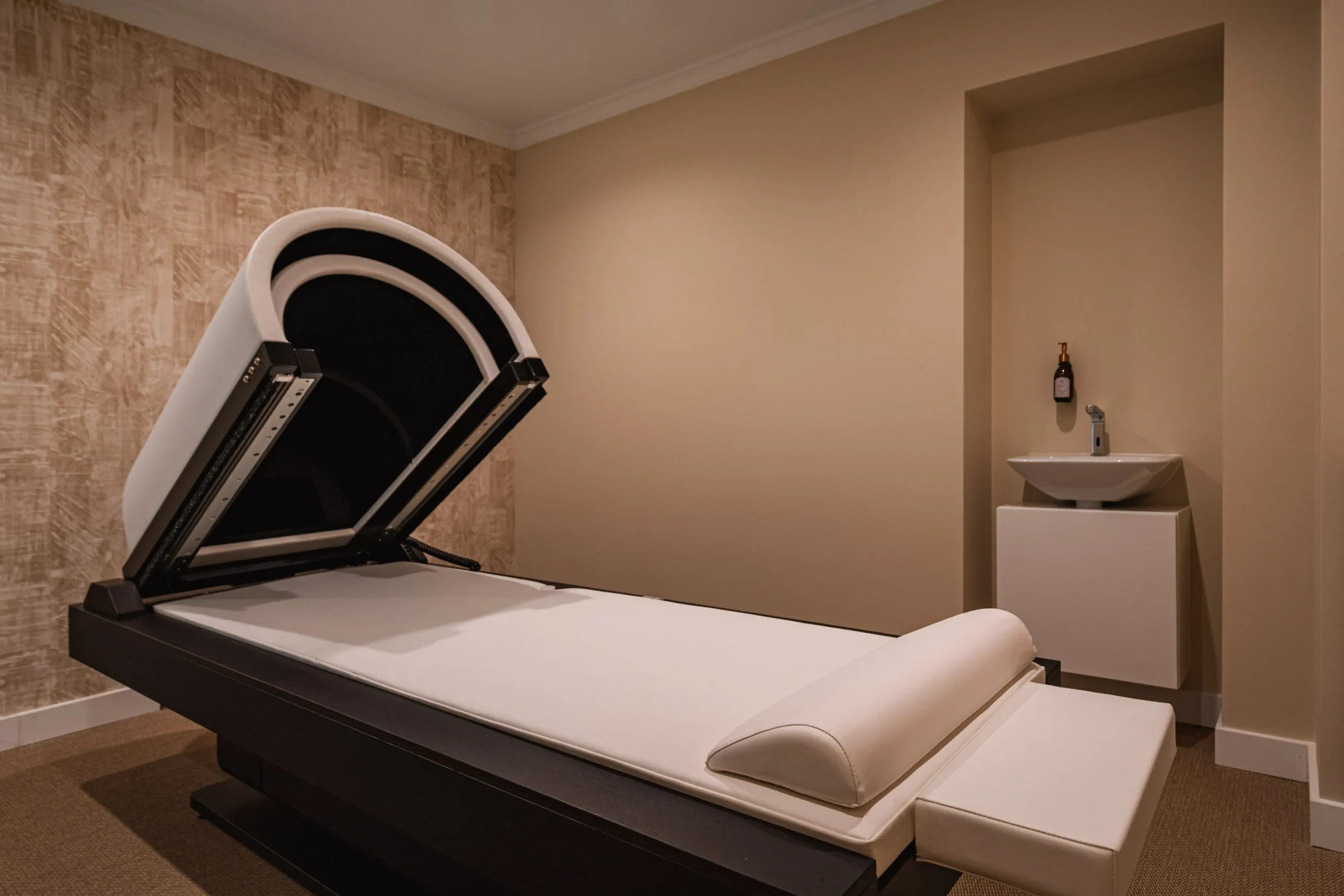 Modern massage room with a high-tech treatment bed and a small sink in a cozy, neutral-toned space.
