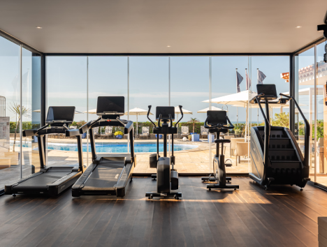 Gym equipment in front of large windows overlooking a pool area with umbrellas.