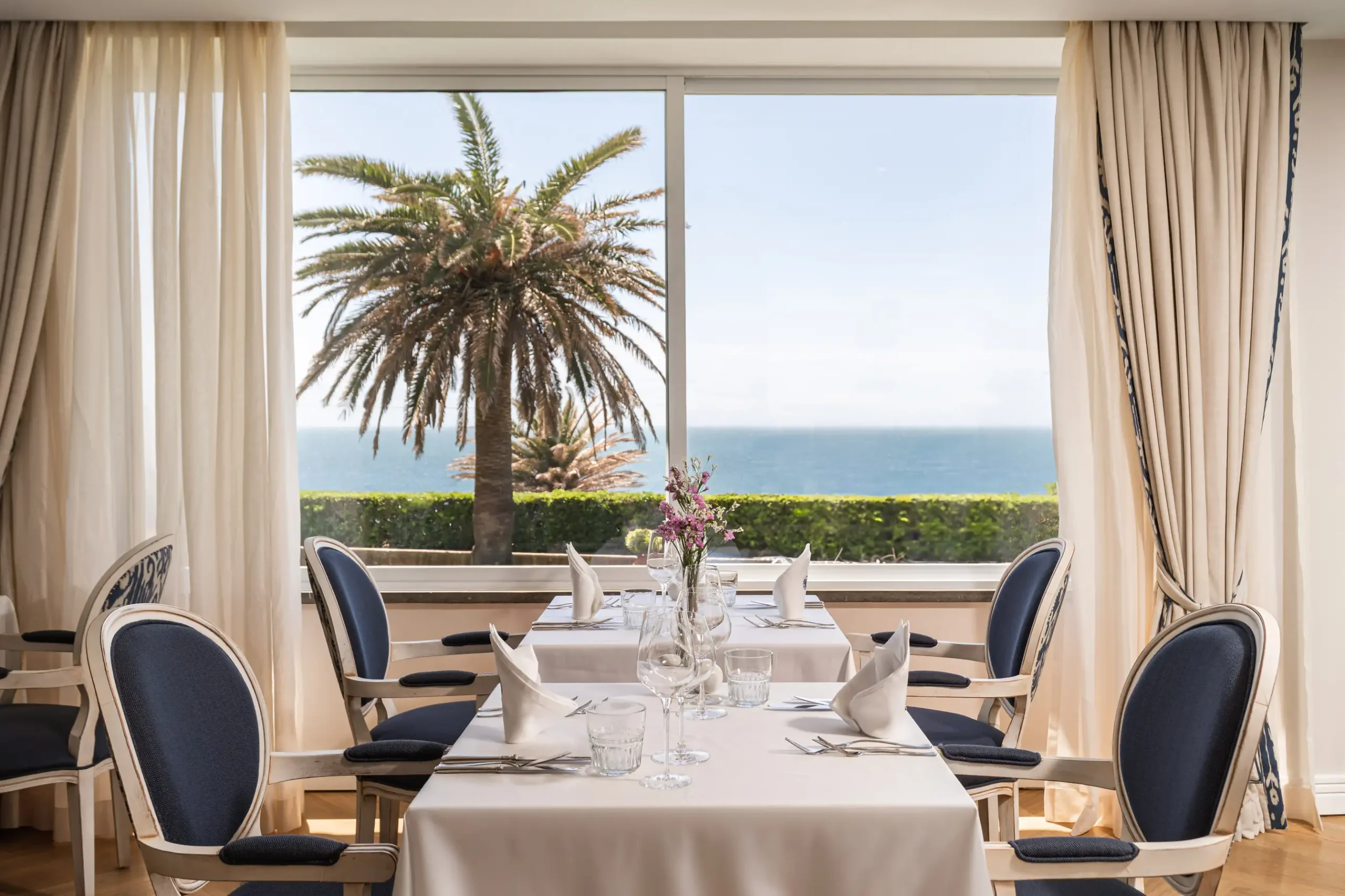 Dining room with a sea view, palm tree, and set tables with white tablecloths and blue chairs.