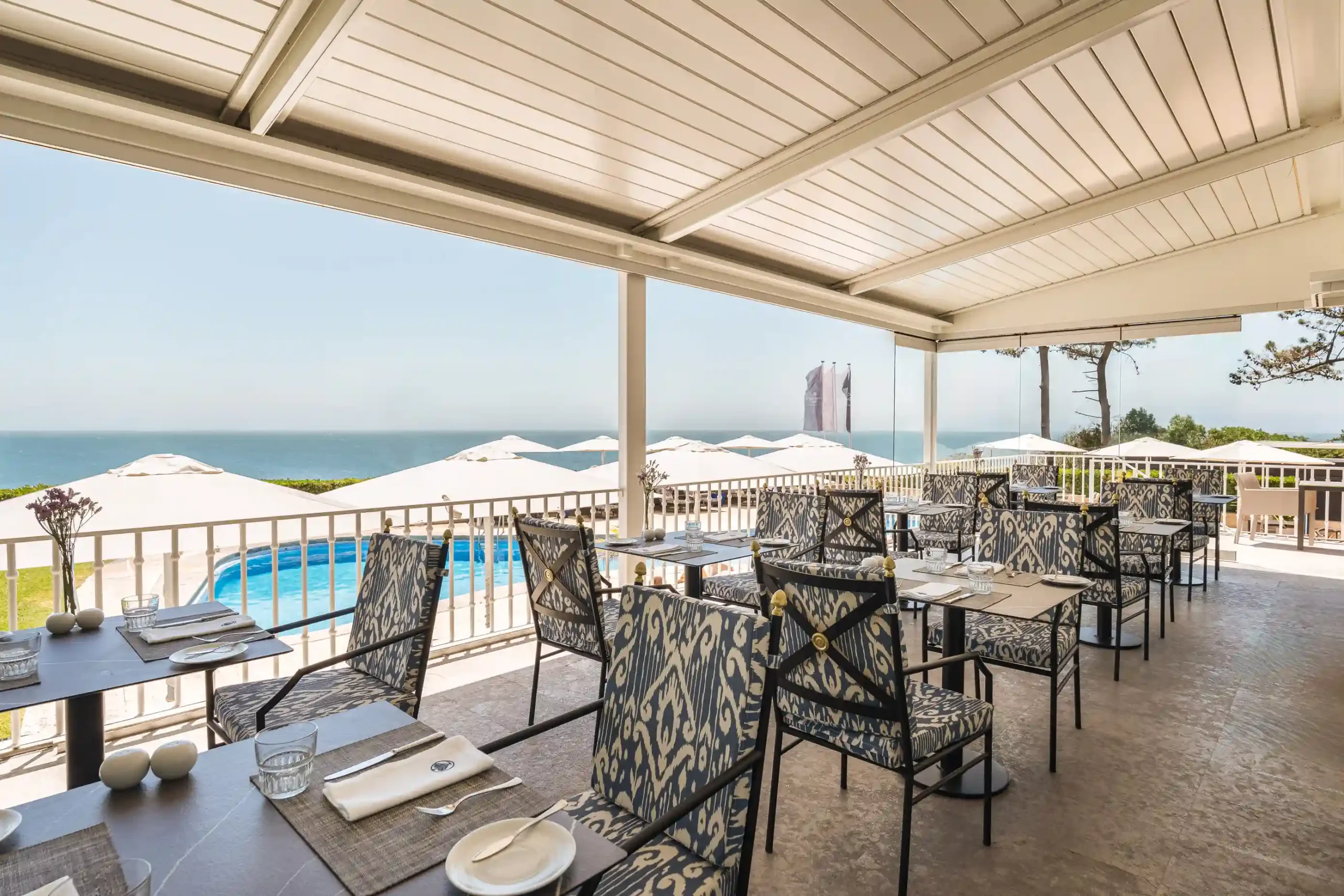 Outdoor dining area with ocean view, patterned chairs, and set tables under a covered patio.