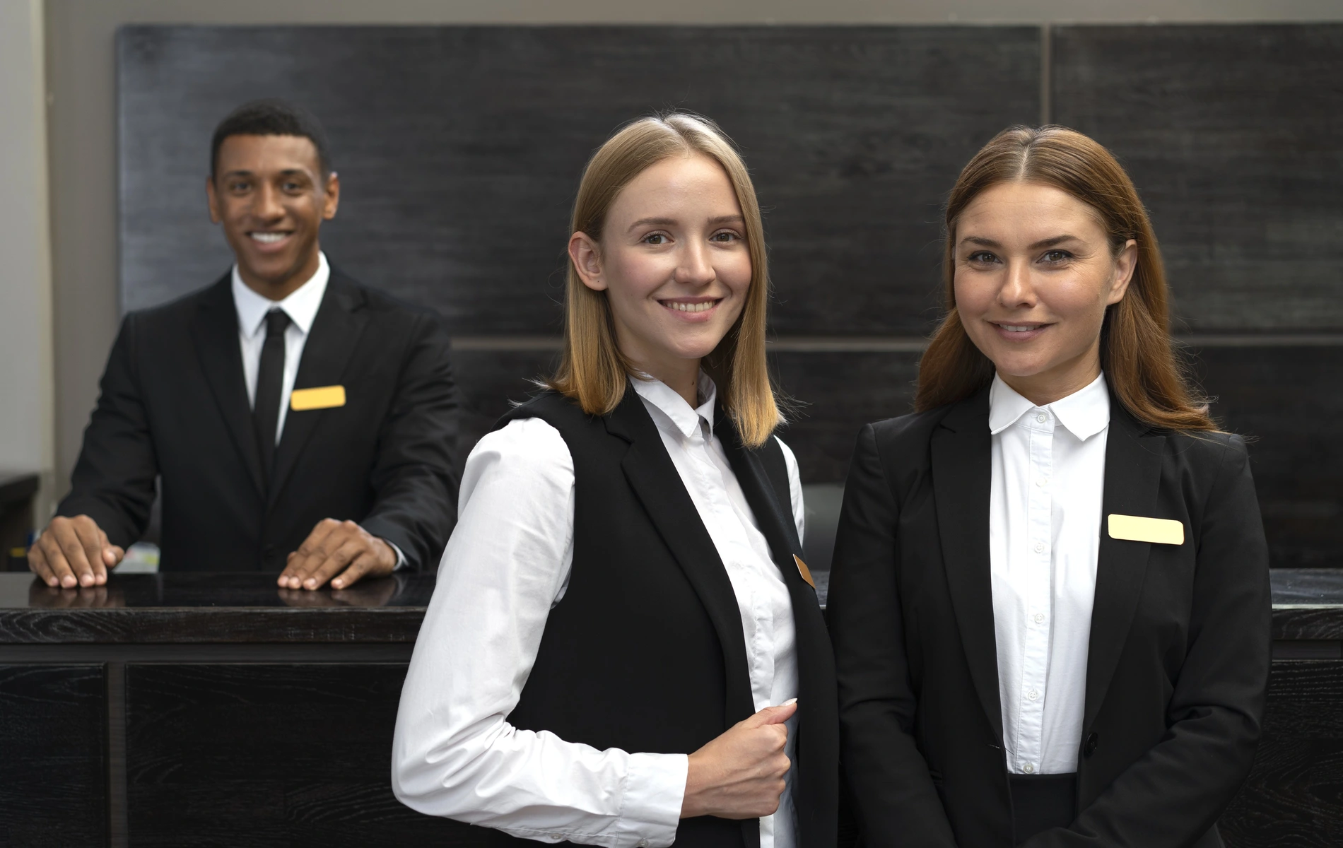 Three hotel receptionists smiling in uniforms behind a dark desk.