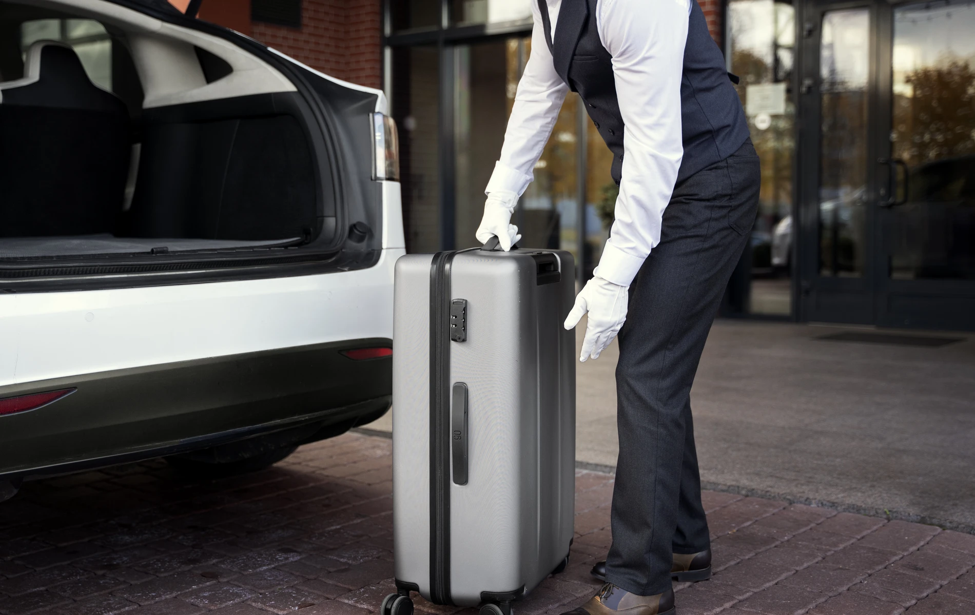 Man with gloves loading a suitcase into a car trunk outside a building.