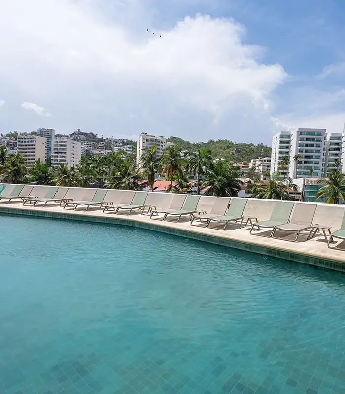 Piscina con reposeras vac&iacute;as, edificios de fondo y palmeras bajo un cielo parcialmente nublado.