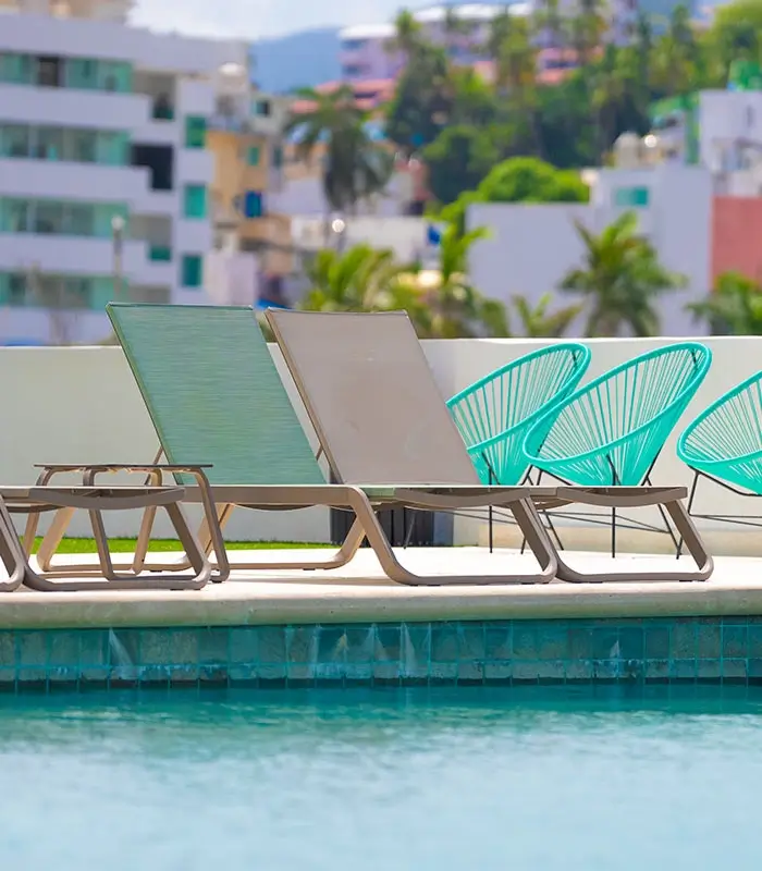 Sillas reclinables y asientos verdes junto a una piscina, con edificios y &aacute;rboles al fondo.