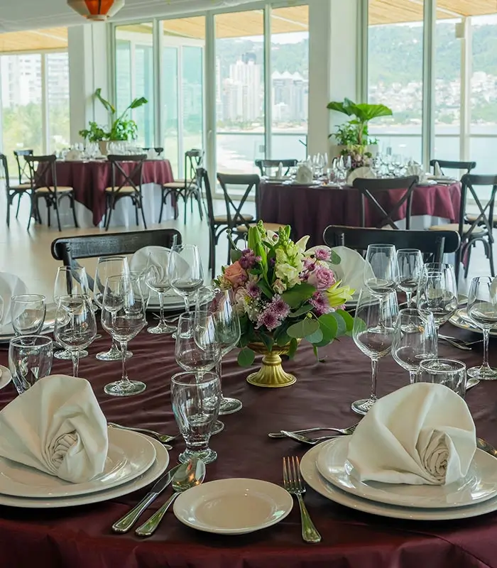 Mesa elegante con manteles burdeos, cubiertos, copas y centro de mesa floral. Vista panor&aacute;mica.