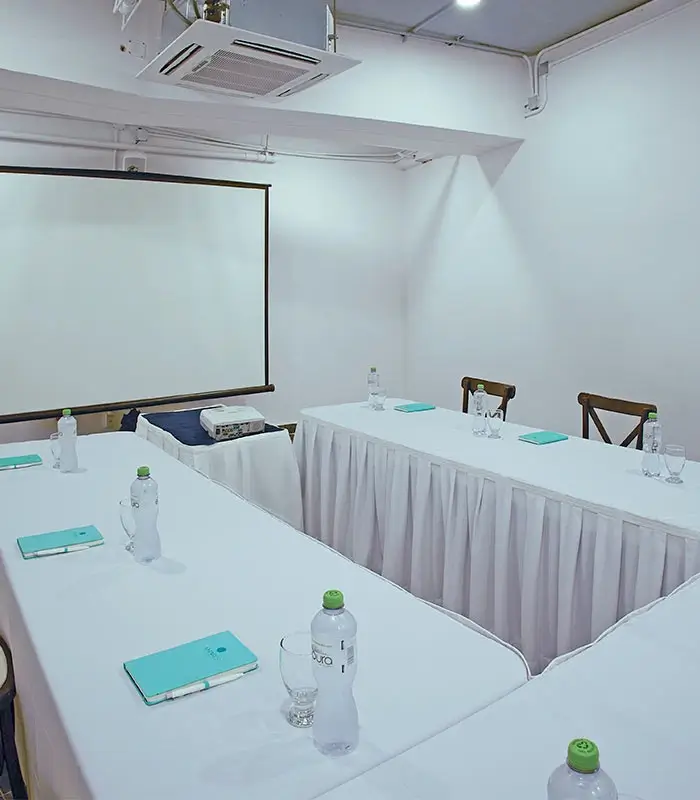Sala de reuniones con mesa en U, proyectores, cuadernos y botellas de agua sobre la mesa.