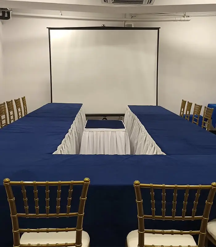 Sala de conferencias con sillas doradas y mesas en forma de "U", listas para evento. Pantalla blanca.
