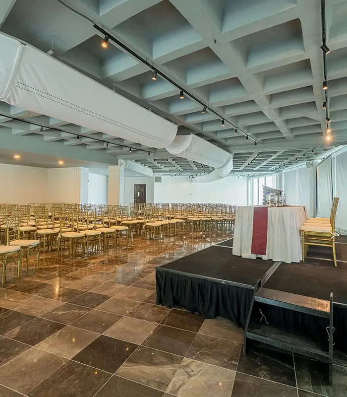 Sala de conferencias con sillas doradas y un podio sobre piso de m&aacute;rmol, decorada con cortinas blancas.
